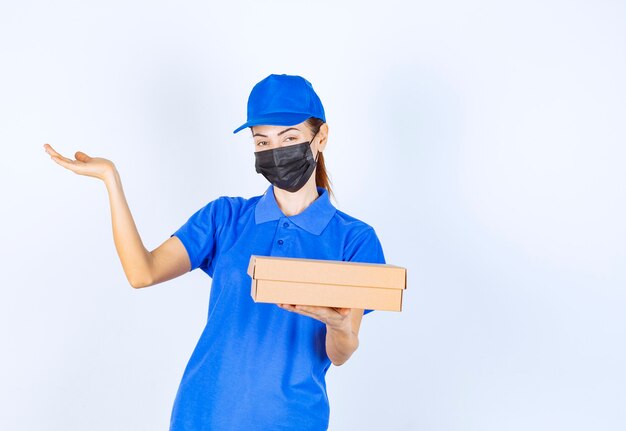 Mensajero mujer en uniforme azul y mascarilla sosteniendo una caja de cartón y apuntando a algún lugar.