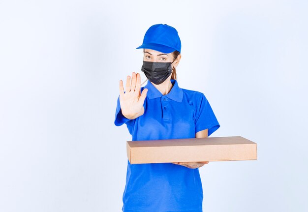 Mensajero mujer en uniforme azul y mascarilla sosteniendo una caja de cartón y deteniendo algo.