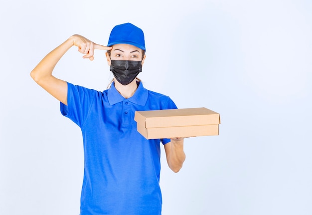 Mensajero mujer en uniforme azul y mascarilla sosteniendo una caja de cartón y pensando en la entrega.