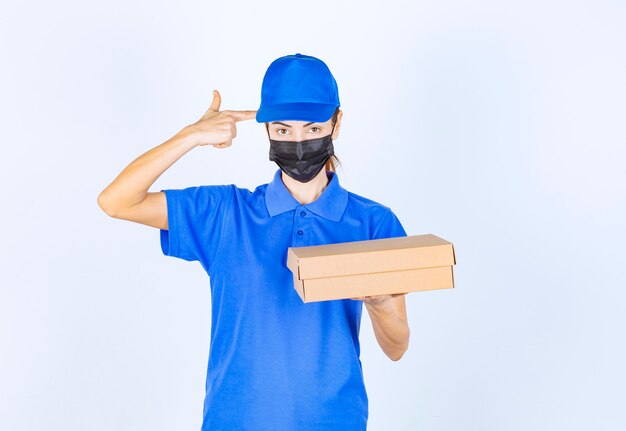 Mensajero mujer en uniforme azul y mascarilla sosteniendo una caja de cartón y pensando en la entrega.