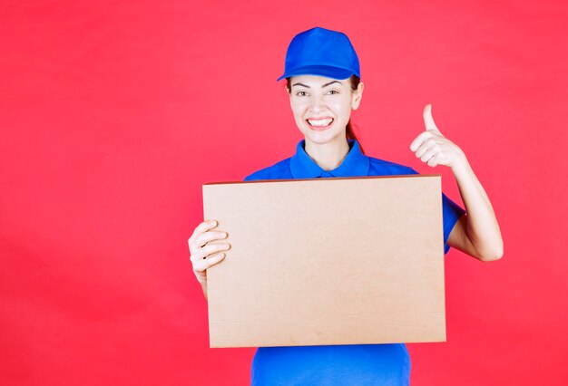 Mensajero mujer en uniforme azul sosteniendo una caja de cartón de pizza para llevar y mostrando el signo de disfrute.