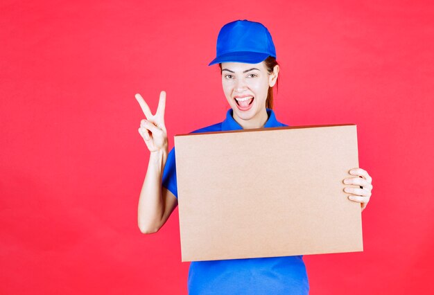 Mensajero mujer en uniforme azul sosteniendo una caja de cartón de pizza para llevar y mostrando el signo de disfrute.