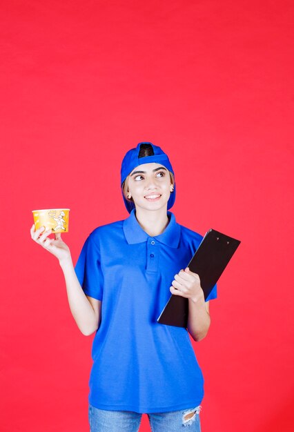 Mensajero mujer en uniforme azul sosteniendo una taza de fideos amarillo y una carpeta negra.