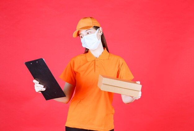 Mensajero mujer en uniforme de color naranja y máscara sosteniendo una caja de cartón y leyendo el nombre y la dirección.