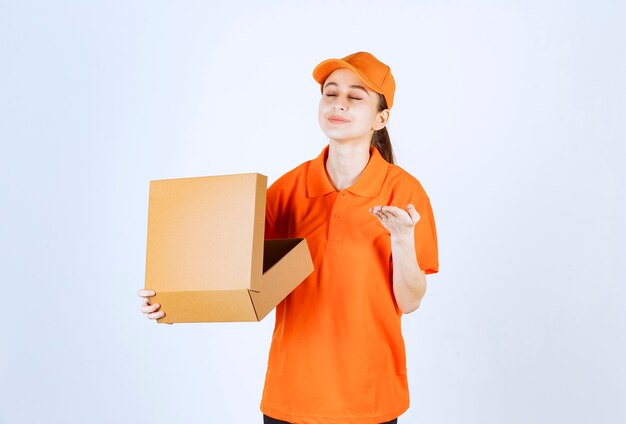 Mensajero mujer en uniforme naranja sosteniendo una caja de cartón abierta y oliendo el producto en el interior.