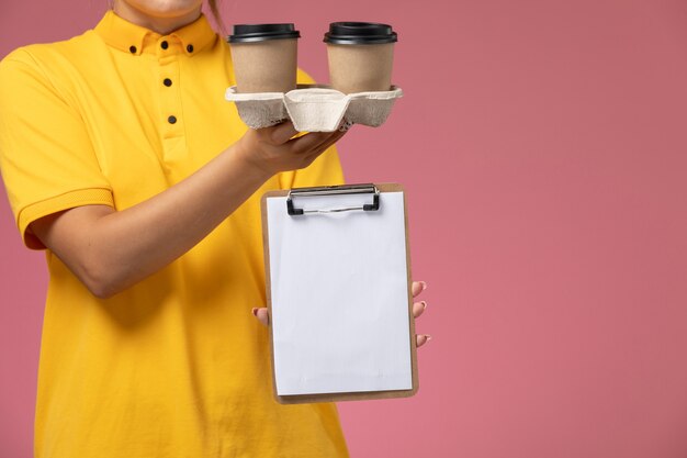 Mensajero mujer vista frontal en capa amarilla uniforme amarillo sosteniendo café y bloc de notas en el trabajo de trabajo de entrega uniforme de fondo rosa