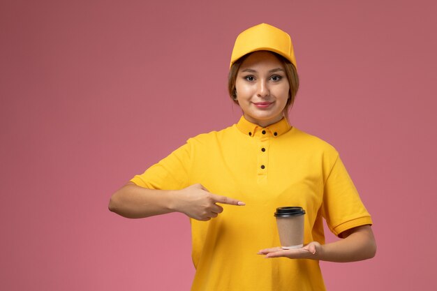 Mensajero mujer vista frontal en capa amarilla uniforme amarillo sosteniendo una taza de café de plástico en el trabajo de trabajo de entrega uniforme de fondo rosa