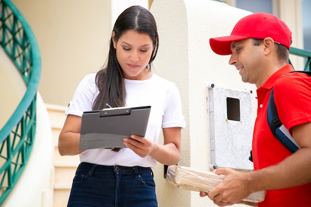 Mensajero positivo en uniforme que entrega el paquete a la puerta del cliente. Mujer firmando para recibir el paquete. Concepto de servicio de envío o entrega