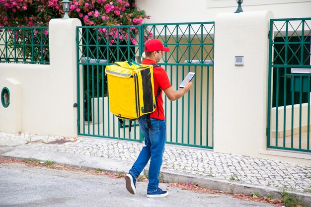 Mensajero con tableta de consultoría de mochila de alimentos isotérmicos, verificando la dirección y caminando hacia la puerta y el timbre. Concepto de servicio de comunicación o entrega