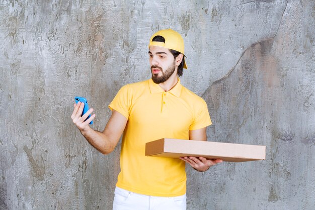 Mensajero con uniforme amarillo sosteniendo una caja de pizza para llevar y hablando por teléfono o haciendo una videollamada.