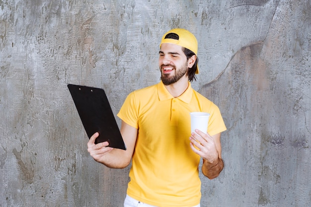 Mensajero en uniforme amarillo sosteniendo un vaso desechable y leyendo la lista de clientes