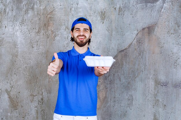 Mensajero en uniforme azul sosteniendo una caja de comida para llevar de plástico y mostrando un signo de mano positivo