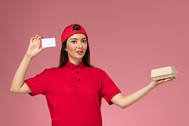 Mensajero de vista frontal con capa de uniforme rojo con pequeño paquete de comida de entrega y tarjeta en sus manos en la pared rosa claro, trabajo de empleado de entrega de servicio