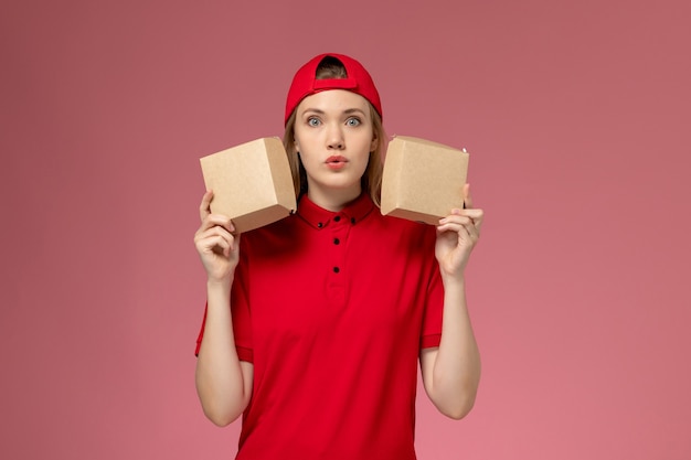 Mensajero de vista frontal en uniforme rojo y capa sosteniendo pequeños paquetes de comida de entrega en la pared rosa claro, trabajador de entrega uniforme de trabajo de servicio