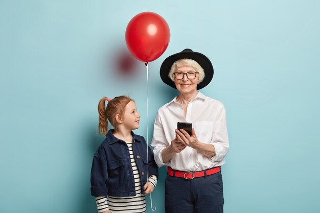 Los mensajes de anciana alegres en el chat en línea, siempre en contacto, viste un atuendo elegante. Atractiva chica pelirroja con cola de caballo, sostiene globo rojo, felicita a la abuela con aniversario de cumpleaños