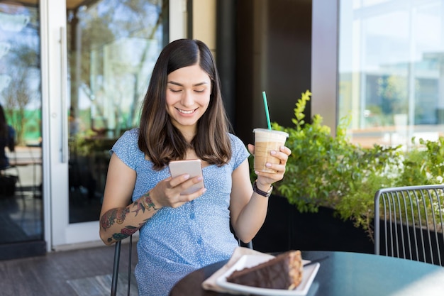 Mensajes de influencia sonrientes en el teléfono móvil mientras toma un café en la cafetería