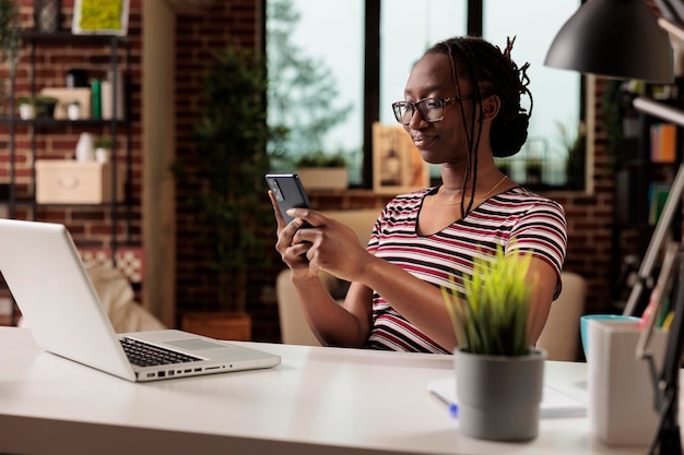 Mensajes de mujer sonriente en el teléfono inteligente, chateando en la red social en el escritorio de la oficina en casa. Empleado con anteojos que tiene un descanso del trabajo, estudiante remoto navegando por Internet en el teléfono móvil