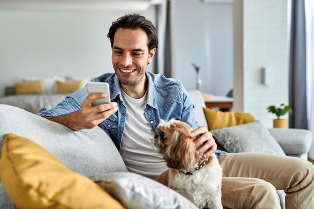 Mensajes de texto de hombre feliz joven en el teléfono celular mientras está sentado en el sofá con su perro