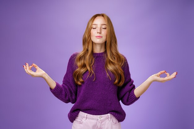 Mente sana en cuerpo. Mujer atractiva pacífica y feliz con cabello rojo y pecas cierra los ojos y sonríe de sentimientos tranquilos y aliviados mientras medita en posición de loto con gesto de mudra, haciendo yoga.