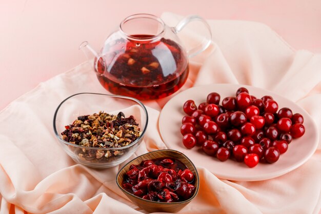 Mermelada de cerezas con cerezas, té, hierbas secas en un tazón sobre textil y rosa