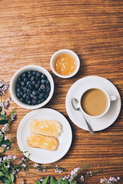 Mermelada extendida sobre una tostada; arándanos; Taza de café y flores rosadas en mesa de madera