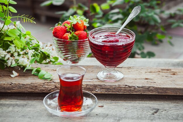 Mermelada de fresa en un plato con cuchara, un vaso de té, fresas, vista desde arriba de la planta en la mesa de madera y pavimento