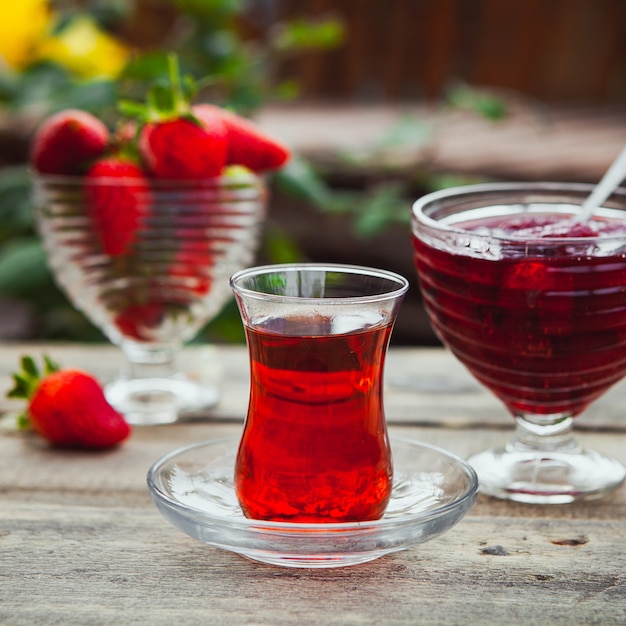 Mermelada de fresa en un plato con un vaso de té, cuchara, fresas, vista lateral en la mesa de madera y patio