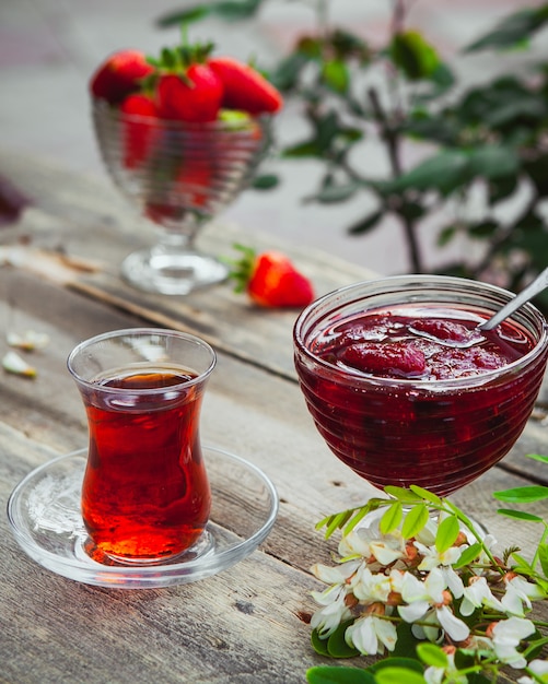 Mermelada de fresa con un vaso de té, cuchara, fresas, plantas en un plato en la mesa de madera y pavimento, vista de ángulo alto.