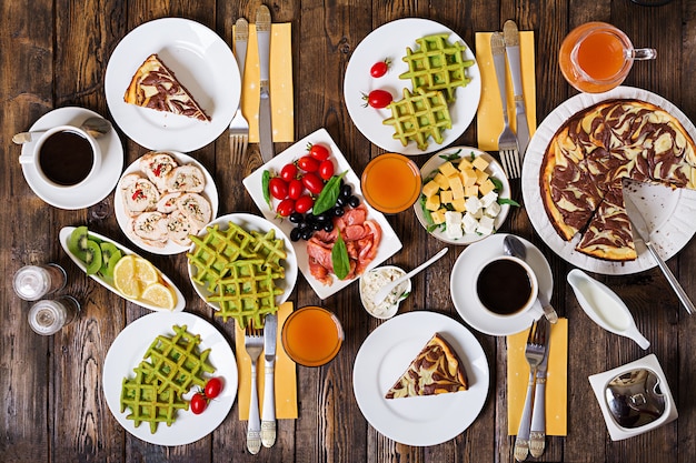 Mesa de comida de desayuno. Brunch festivo, variedad de comidas con waffles de espinacas, salmón, queso, aceitunas, rollitos de pollo y tarta de queso. Vista superior. Lay Flat