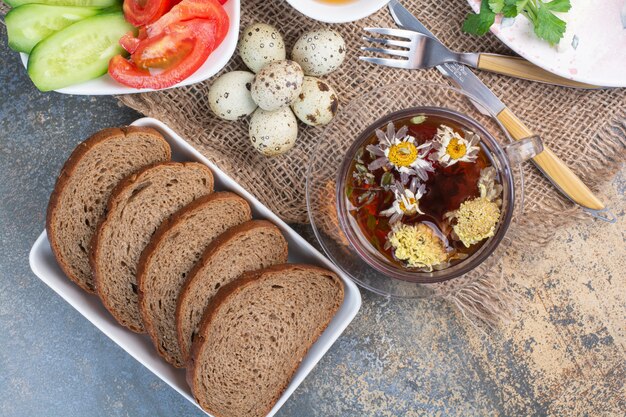 Mesa de desayuno con verduras, té, pan y huevos sobre arpillera.