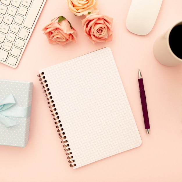 Mesa de escritorio con rosas rosadas, taza de café, cuaderno espiral en blanco, bolígrafo. Vista superior, plano