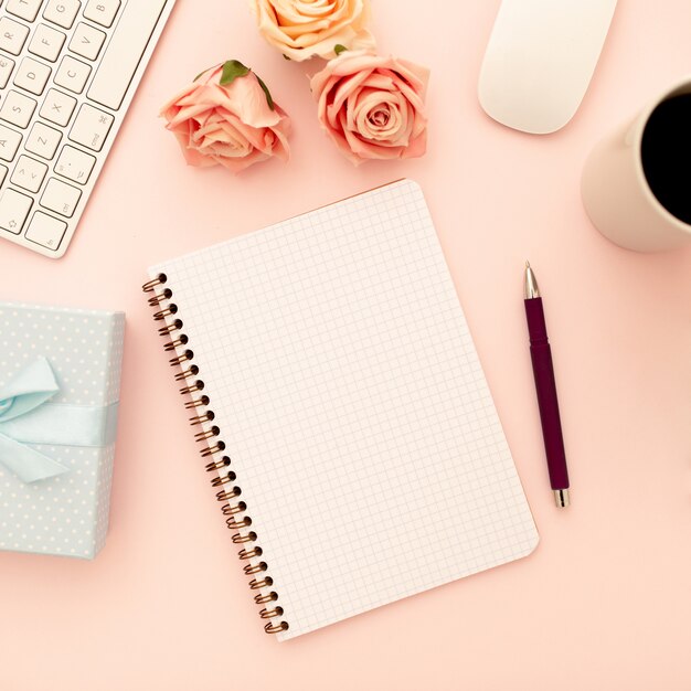 Mesa de escritorio con rosas rosadas, taza de café, cuaderno espiral en blanco, bolígrafo. Vista superior, plano