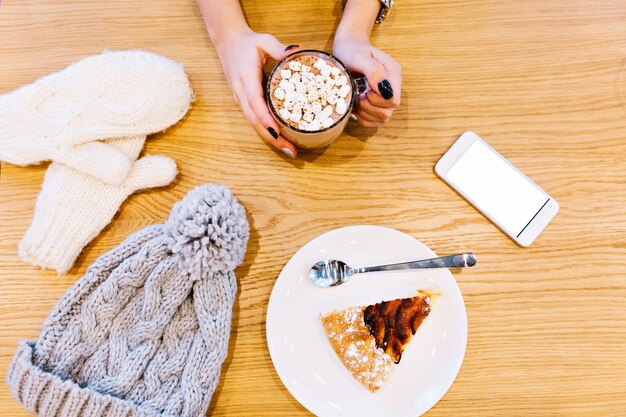 Mesa con guantes blancos de invierno, teléfono, chocolate caliente por niña y trozo de tarta, gorro gris tejido.