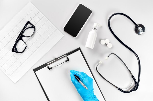 Mesa del médico, herramientas, instrumentos médicos, estetoscopio, trabajo en el hospital sobre fondo blanco plano. La mano del médico en un guante azul registra los indicadores en un papel blanco en blanco. Vista superior. Quieto