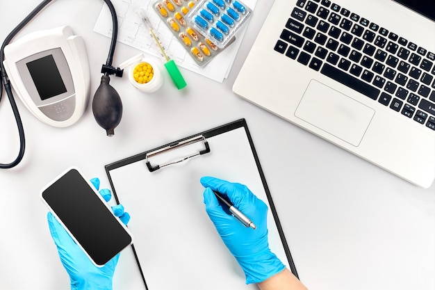 Mesa del médico, herramientas, instrumentos médicos, tonómetro de terapeuta, presión arterial, trabajo en el hospital sobre fondo blanco plano. La mano del doctor en un guante azul registra los indicadores en un papel blanco.