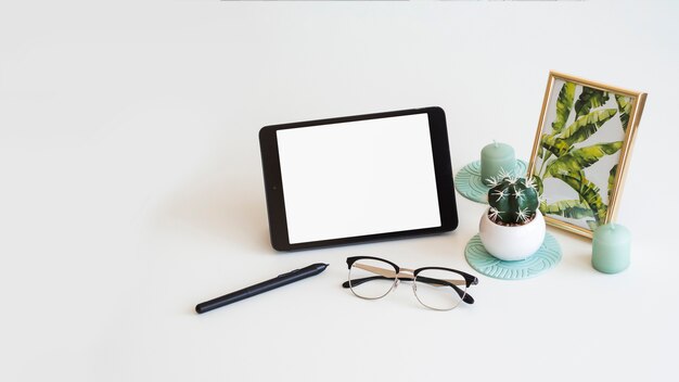 Mesa con tableta cerca de marco de fotos, cactus, pluma y lentes.