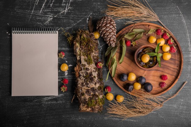 Mezcla de bayas en una bandeja de madera con ramas de roble, conos y un cuaderno a un lado