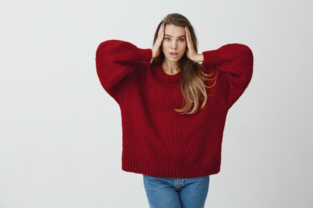 Mi cabeza está dando vueltas. Retrato de mujer sensual expresiva con apariencia atractiva de pie en suéter rojo suelto, cogidos de la mano en la cara y haciendo expresión de modelo