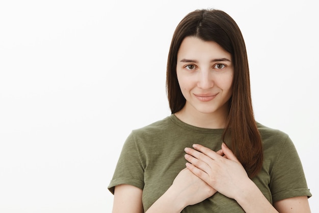 Mi corazón está contigo. Romántica tierna y sensual mujer morena en camiseta verde oliva cogidos de la mano en el pecho y sonriendo despreocupada expresando romance y actitud de afecto sobre pared gris