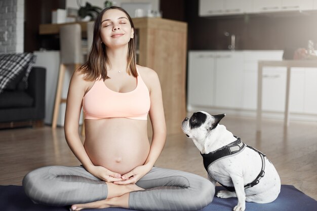 Mi entrenador de cuatro patas me vigila. Filmación en interiores de atractiva y relajada mujer europea embarazada en elegante ropa deportiva, sonriendo alegremente mientras está sentado en pose de yoga en la almohadilla de rollo en casa, meditando