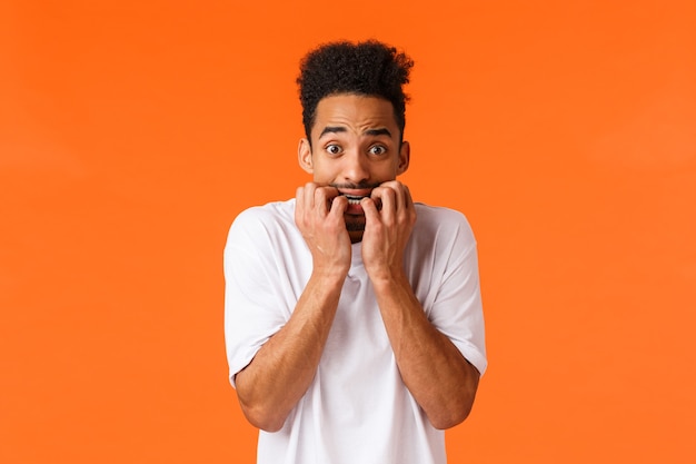 Miedo, películas de terror, concepto de emociones. Asustado e inseguro, joven tímido hipster hombre afroamericano, mordiéndose las uñas, mirando a la cámara ansioso, asustado de que alguien sepa, fondo naranja