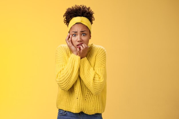 Miedo preocupado nervioso joven empleada afroamericana preocupado ansioso ser despedido apretar los dientes agacharse tocar cara inseguro poco dedos de pie asustado aterrorizado, fondo amarillo.