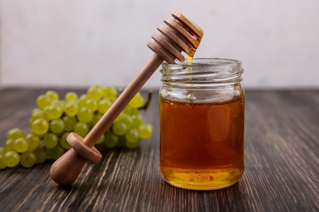 Miel de vista frontal en un frasco con una cuchara de madera y uvas verdes y sobre un fondo de madera