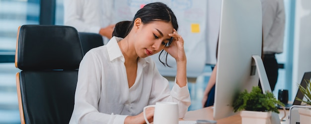 Milenaria joven empresaria china trabajando estresarse con el problema de investigación del proyecto en el escritorio de la computadora en la sala de reuniones en la pequeña oficina moderna Concepto de síndrome de burnout ocupacional de personas de Asia.