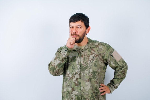 El militar está pensando sosteniendo el dedo índice en la mejilla y poniendo la otra mano en la cintura sobre fondo blanco.