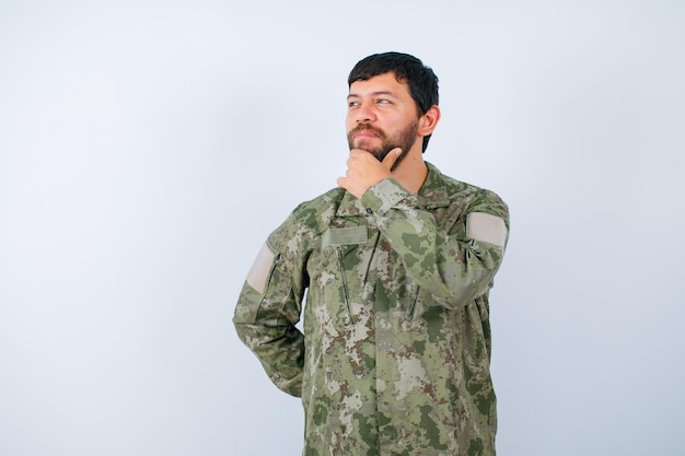 El militar está pensando sosteniendo la mano en la barbilla y poniendo la otra mano en la cintura sobre fondo blanco.