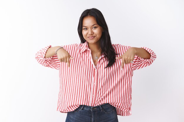 Mira esto ahora. Retrato de mujer vietnamita atractiva intrigante en blusa de moda a rayas apuntando hacia abajo y mirando con sonrisa a la cámara que muestra un espacio de copia interesante sobre la pared blanca