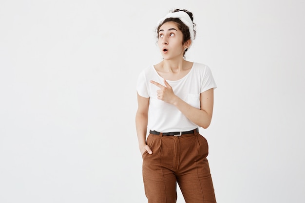 ¡Mira esto! La alegre y joven mujer de cabello oscuro con moño hizo estallar los ojos con entusiasmo, alejando su dedo índice, indicando espacio de copia en la pared blanca en blanco para información promocional