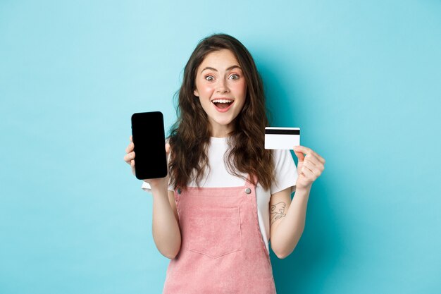 Mira aquí. La muchacha linda joven emocionada que muestra la pantalla del móvil vacía y la tarjeta de crédito plástica, mira asombrada a la cámara, colocándose sobre fondo azul.