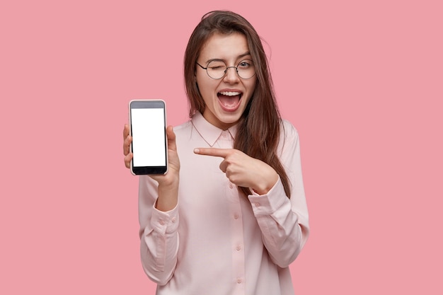 Foto gratuita ¡mira este celular! la mujer feliz y complacida parpadea los ojos, señala con el dedo índice a la pantalla en blanco, muestra un dispositivo moderno